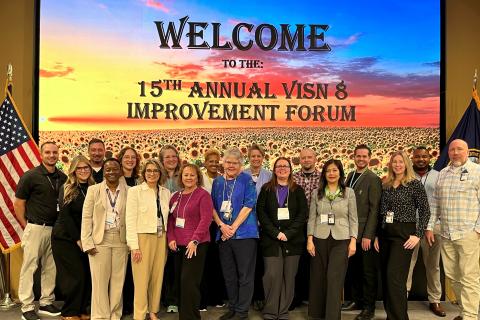 Group of people standing in front of a stage with a sign that says "WELCOME TO THE 15TH ANNUAL VISH & IMPROVEMENT FORUM".
