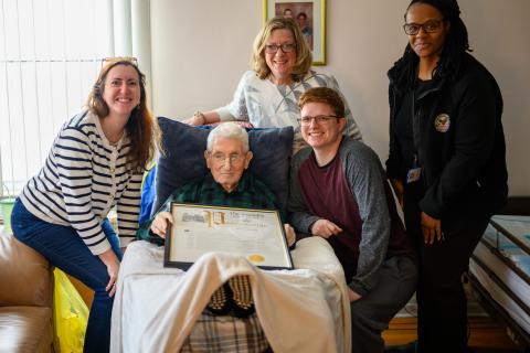 Five people in a living room, one is an elderly man in a chair with a plaque.