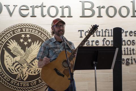 Man with beard and glasses holding guitar in front of Overton Brooks VA Medical Center sign.