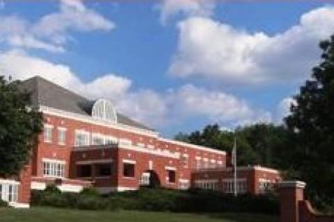 Large red building with many windows and a gray roof, surrounded by trees and grass.