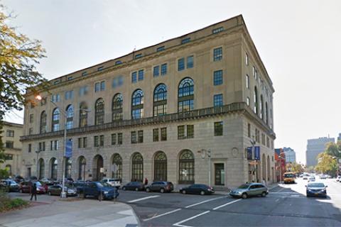 Beige building with arched windows on the first and third floor. Across the street from a tree-lined park 