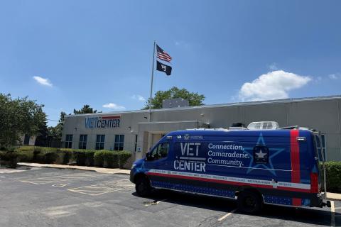 Blue and red van with "VET CENTER" on side parked in front of building with flags.