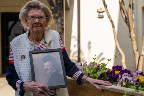 Margaret Brewer, U.S. Navy Veteran, poses in a flower garden at Somerby Baxter Village, in Fort Mill, SC, holding a photo of her in uniform. 