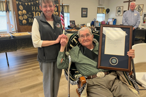 A woman stands next to a seated man holding a framed certificate.
