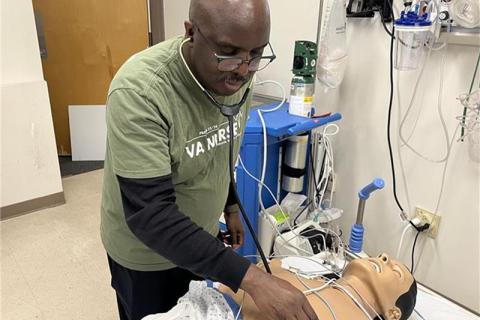 Man in green shirt and black pants using stethoscope on CPR mannequin in hospital room.