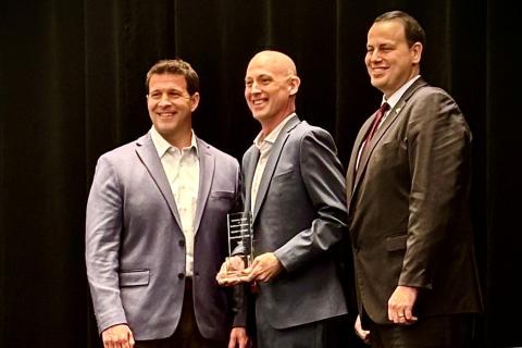 Three men in suits stand on a stage; the middle man holds an award.