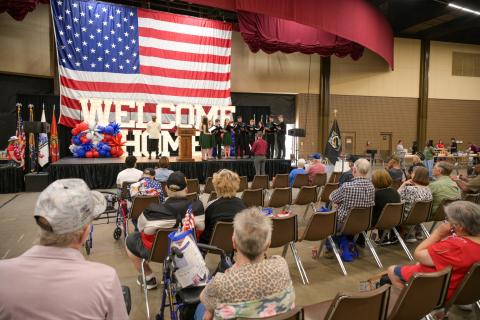 A crowd of people in a large hall with an American flag and "WELCOME HOME" banner.