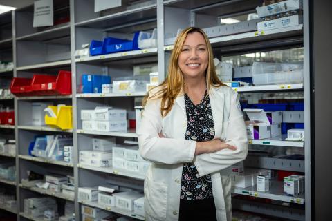 Smiling woman in white lab coat stands with arms crossed in front of shelves full of medicine.