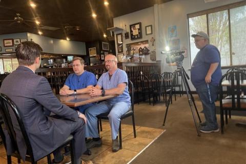 Three men in a restaurant sitting at a table with a camera man recording them.