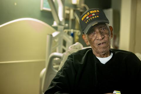 An elderly man in a World War II veteran hat sits in a hospital bed.