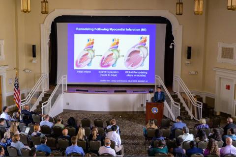 Large lecture hall with people seated facing a stage with a large screen displaying a medical diagram.