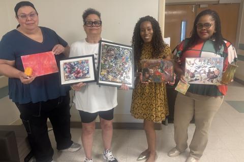 Four women in a hallway display their artwork.