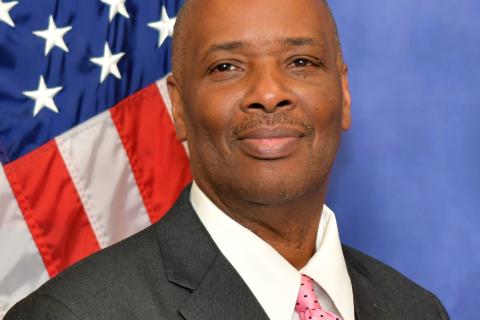 Man in suit and pink tie stands in front of American flag.