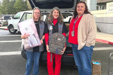 Three women in scrubs holding a mattress and backpack next to a van.