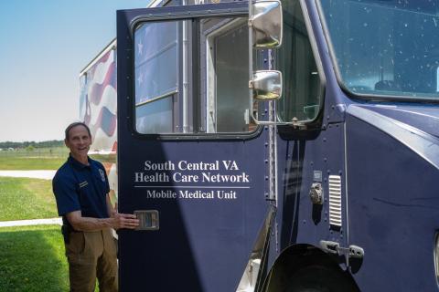 Man in blue shirt and brown pants standing by blue mobile medical unit.