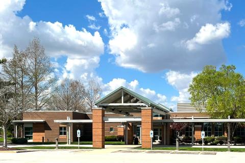 Modern building entrance with covered walkway, trees, and blue sky with clouds.