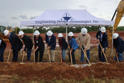 Several people in hard hats stand holding shovels under a tent at a construction site.
