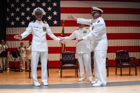 Two navy officers shake hands on stage in front of an American flag.