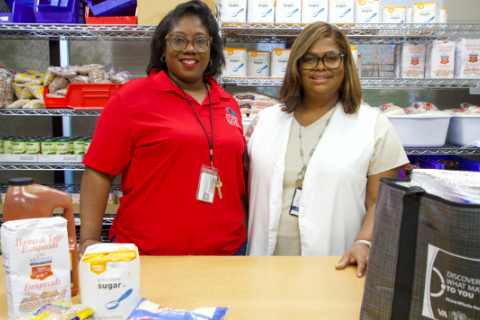 Two women stand behind a table with food items, smiling.