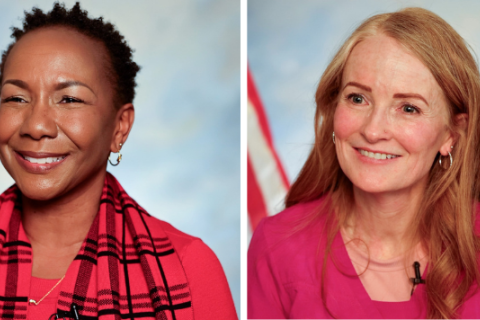 Two smiling women in red and pink shirts with American flags in background.