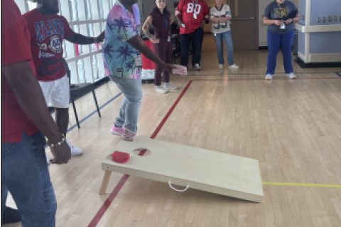 Man in tie-dye shirt throws beanbag onto cornhole board during game in gym.