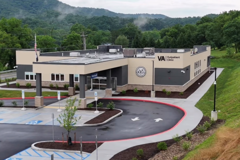 Modern building with circular entrance, clock logo, and parking lot, set against a backdrop of lush greenery and hills.