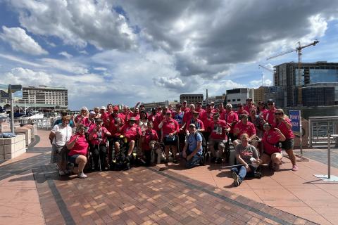 Group of people in wheelchairs and standing, wearing pink shirts, pose on a brick plaza.