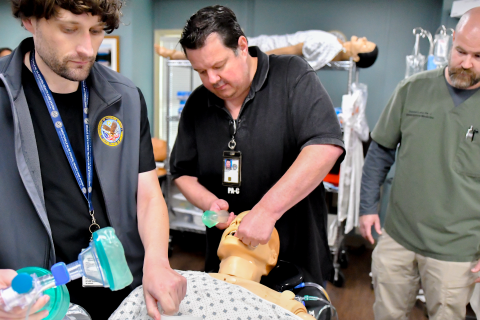 Three men practicing CPR on a dummy in a hospital room.