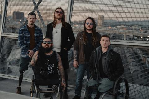 Five men, two in wheelchairs, pose on a bridge with city skyline in background.