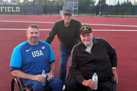 Three men, two in wheelchairs, on a track field.