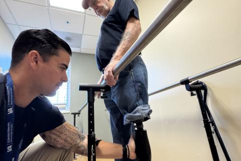 Two men in a hospital room, one helping the other walk with a prosthetic leg.