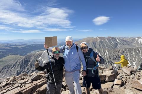 Three hikers on mountain peak, one holding sign, with scenic view.