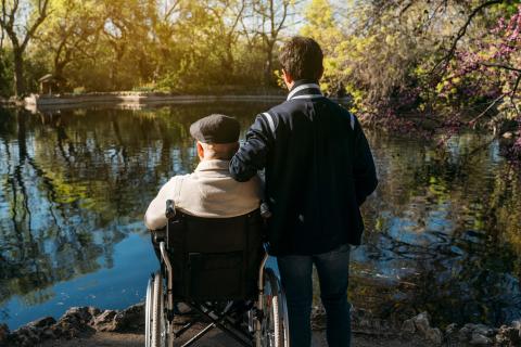 A younger man supports an older man in a wheelchair by a pond.