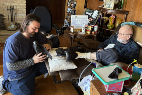 Two men in a living room, one putting on the other's shoe.