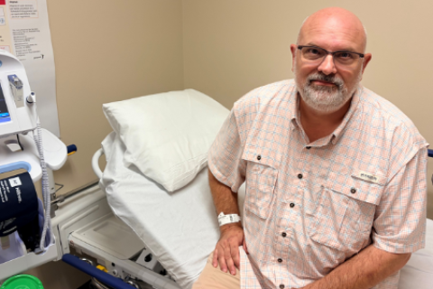 Man with glasses and a beard sitting on a hospital bed next to a monitor.