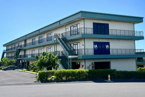 Two-story building with balconies, blue accents, and a parking lot in front.