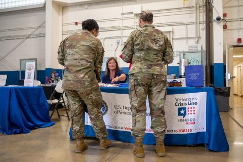 Two soldiers in camouflage uniforms stand facing a table with a woman, indoors.