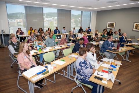 Diverse group of people seated at tables in a conference room with large windows.
