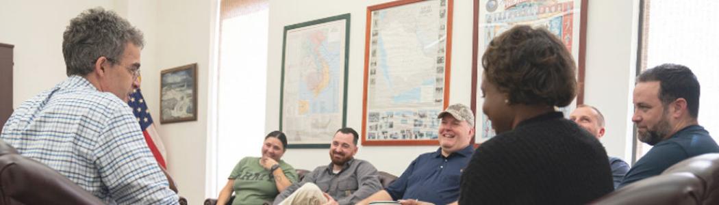 Small group of diverse Veterans talking and smiling with a Vet center counselor, American flag in the background.