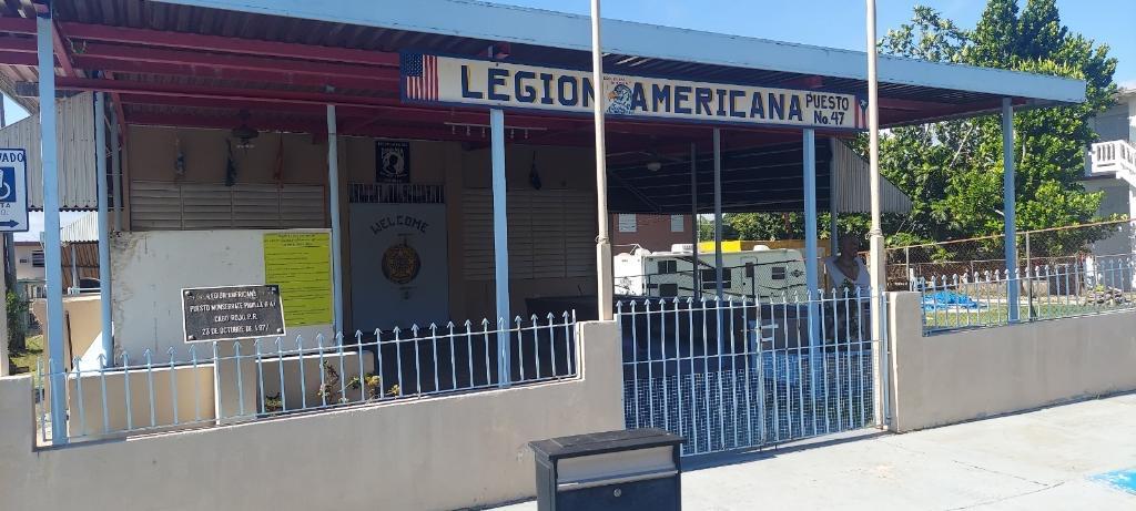 One story building, painted light cream with blue trims on roof, and a cream-colored fence at entrance. Building is identified with a sign on the roof that reads "Legion Americana"
