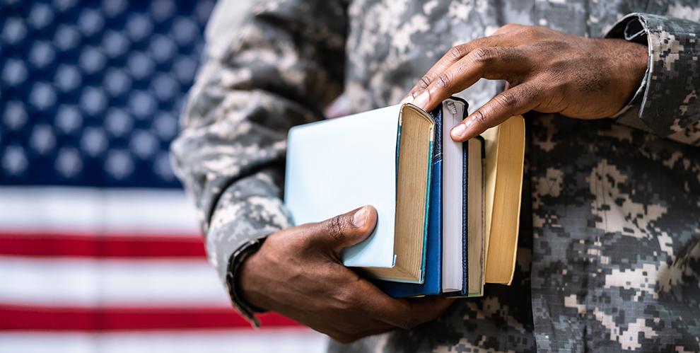 A uniformed servicemember holds a stack of books against an American flag background.