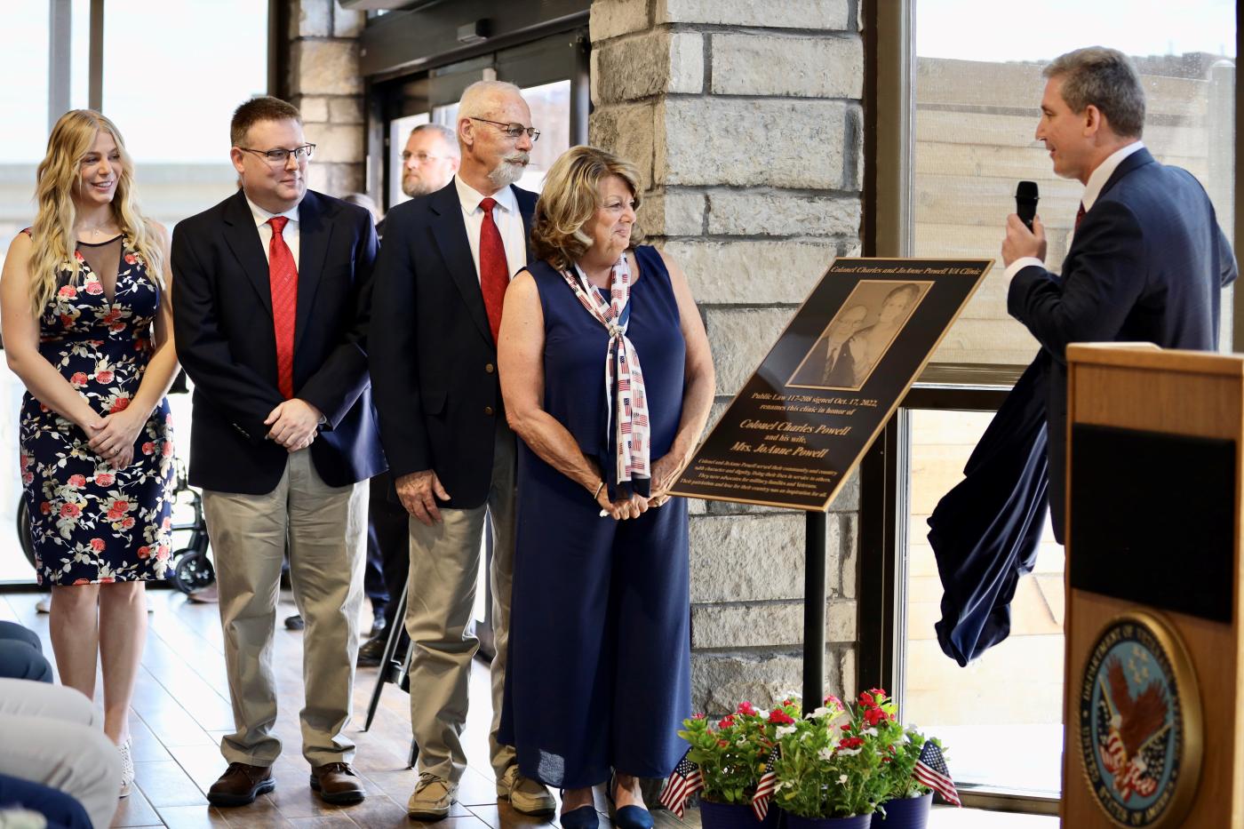 Four people in business attire on the left and a man in a suit on the right looking at a bronze plaque on a stand in the center