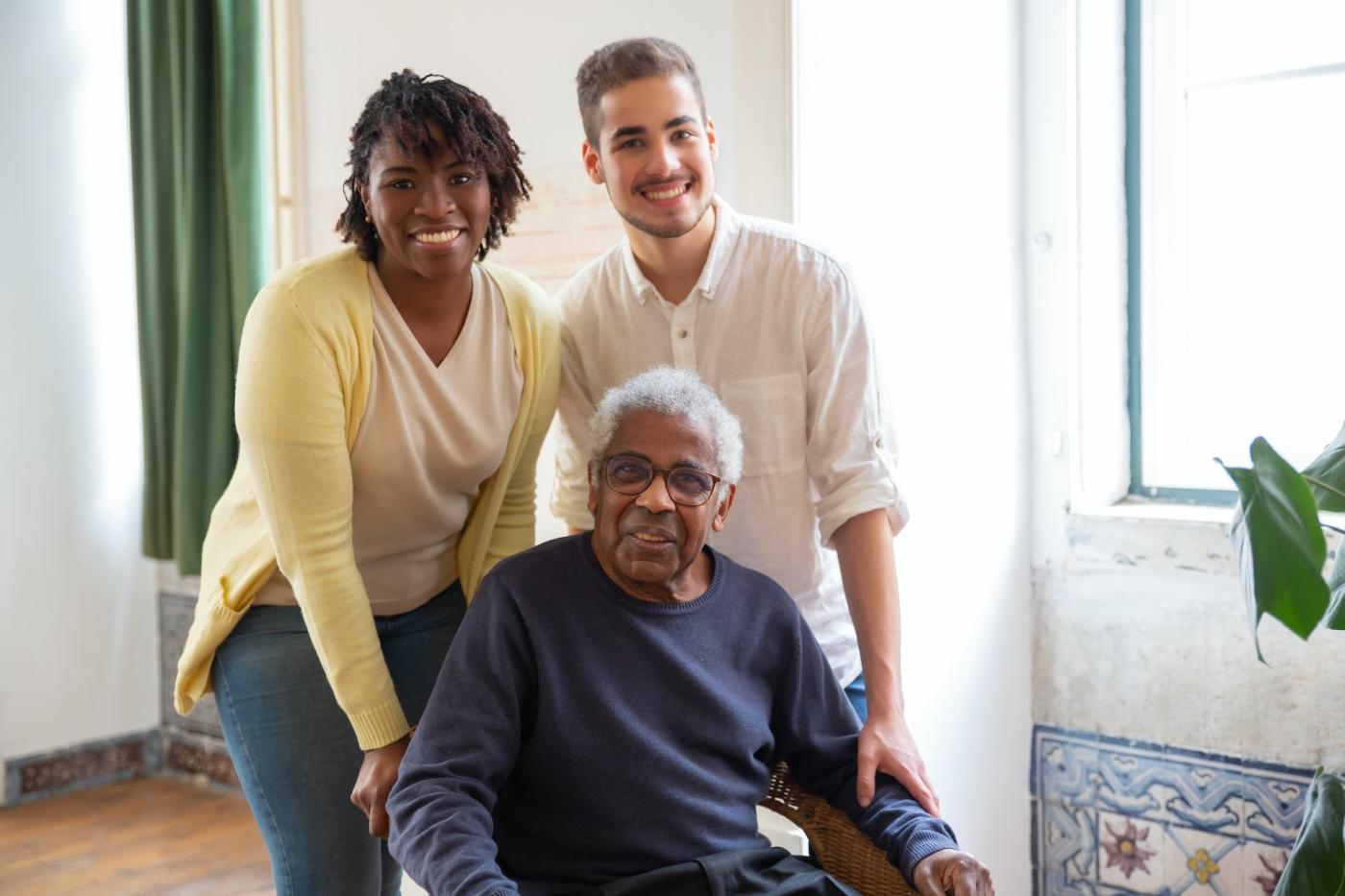 A seated elderly man in front of two standing younger caregivers, all are smiling.