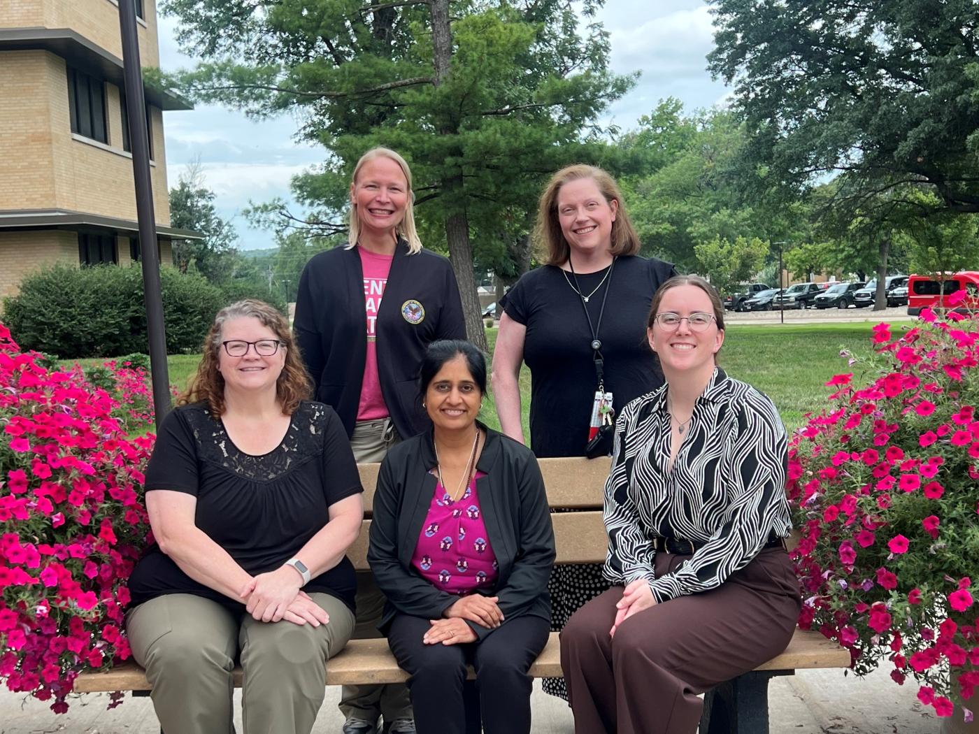 Six women sit on a park bench outside a building.