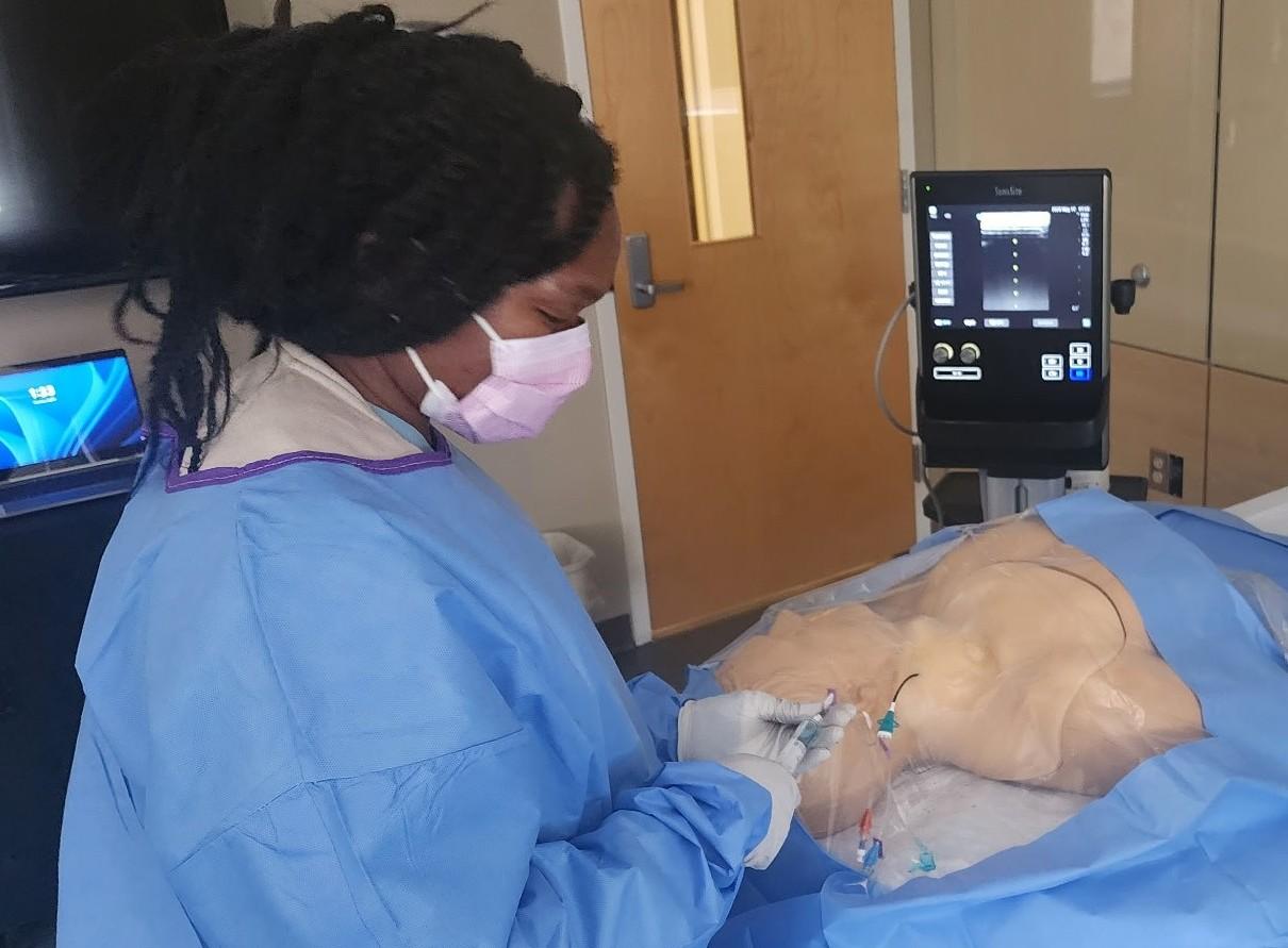 Nurse in blue scrubs practices procedure on a mannequin in a hospital room.