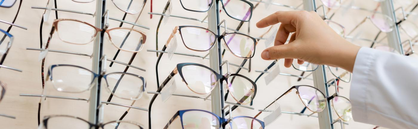 Hand in white lab coat reaching for glasses from display of many different frames.